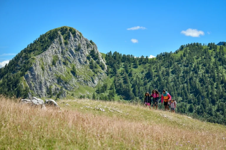 Un gruppo cammina nei prati estivi sotto le pareti rocciose e i boschi delle Alpi Liguri