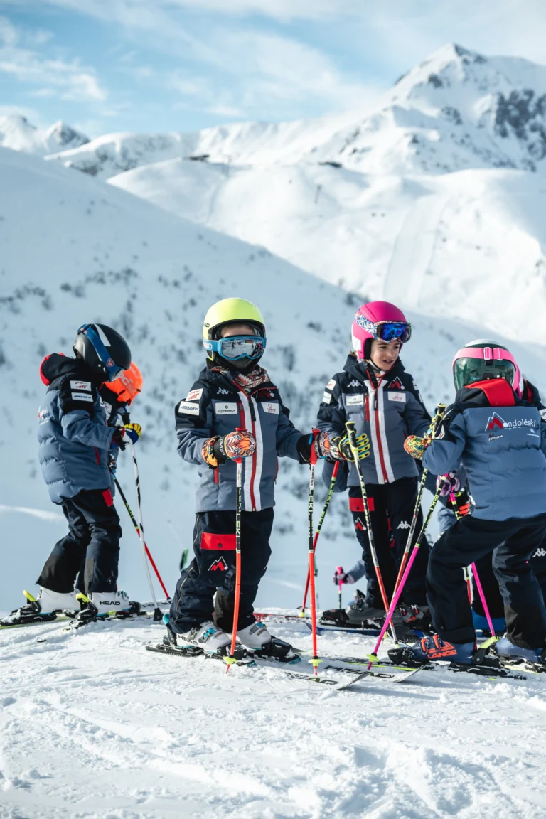 Quattro giovani con sci, caschi e bastoncini davanti alle montagne.