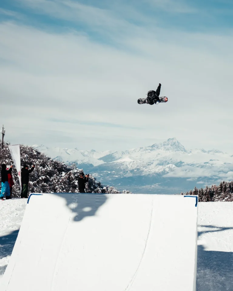 Uno snowboarder in volo sopra un grande salto dello Snowpark, con le Alpi sullo sfondo