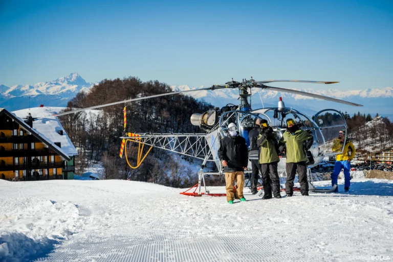 Un gruppo sulla neve accanto a un elicottero fermo, con le montagne sullo sfondo