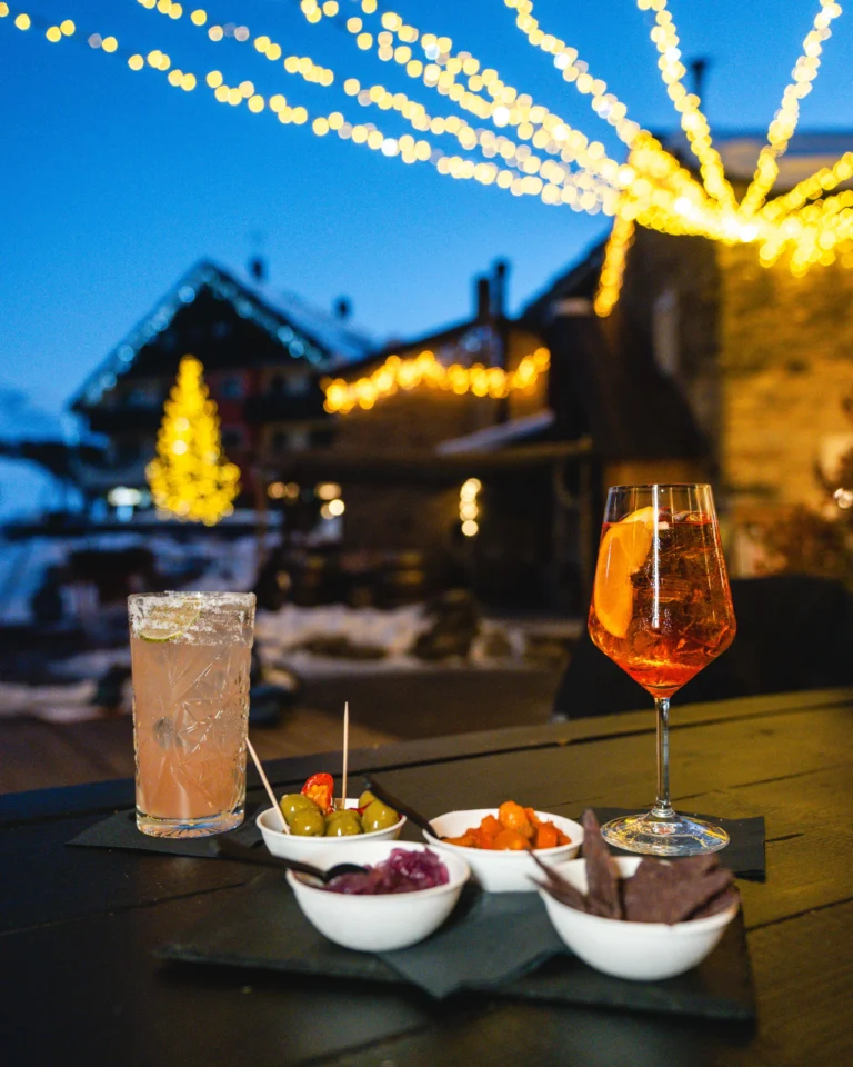 Due cocktail con stuzzichini su un tavolo all’aperto, con luci decorative e chalet sullo sfondo.