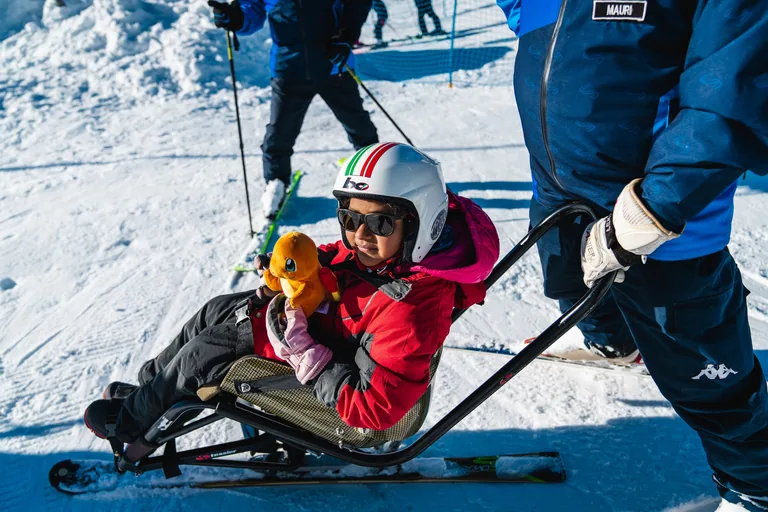 Bambina con casco e occhiali su una seduta da sci adattato, con un pupazzo arancione