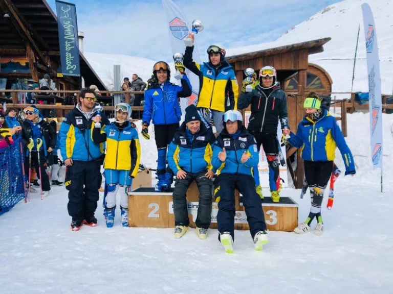 Giovani atleti e tecnici in giacca blu e gialla sul podio davanti a uno chalet, uno di loro alza la coppa