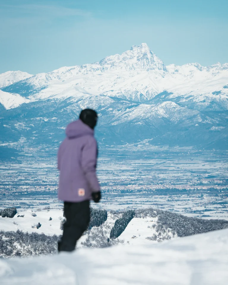 Persona in giacca lilla sul pendio bianco guarda le montagne lontane.