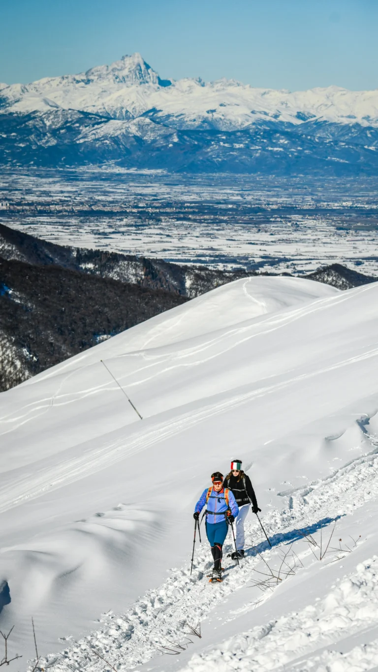 Due escursionisti con ciaspole e bastoncini risalgono un sentiero innevato, con il panorama delle Alpi alle spalle.