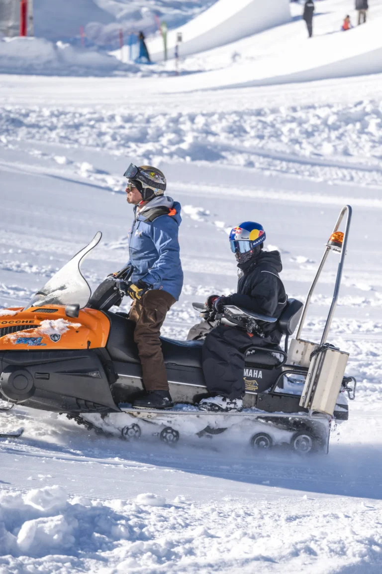 Due persone con casco su una motoslitta nel park, con uno snowboard appoggiato al mezzo