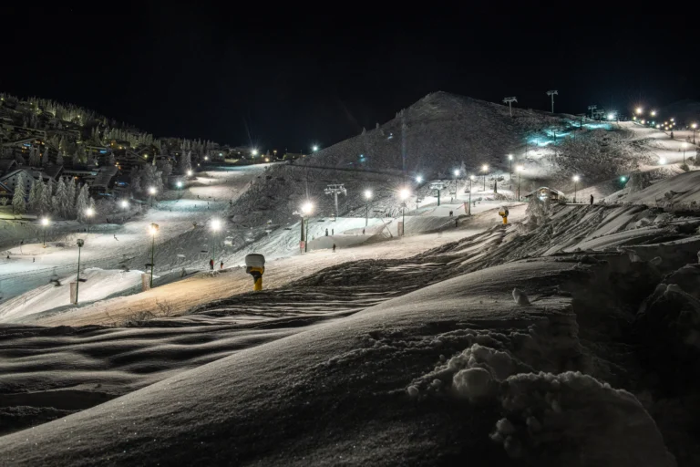 Piste da sci illuminate nella notte, piccoli sciatori e neve ondulata in primo piano.