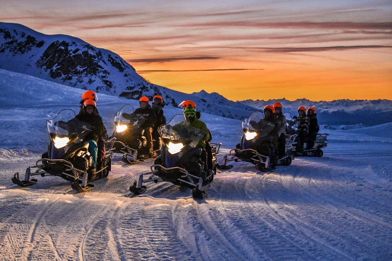 Gruppo con vere motoslitte e fari accesi, davanti alle montagne al tramonto