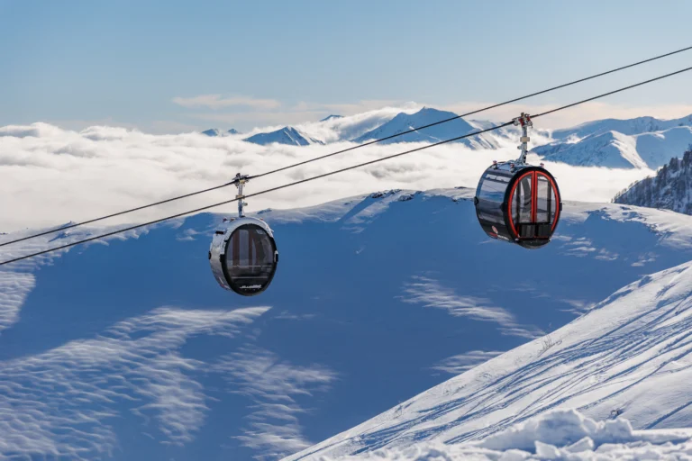 Due cabine della telecabina La Rossa sopra i pendii innevati, con un mare di nuvole e le Alpi sullo sfondo.