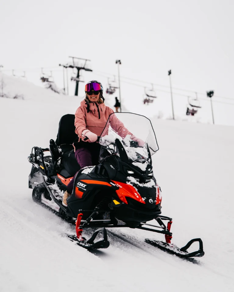 Donna sorridente con casco e giacca rosa alla guida di una motoslitta nera e arancione sulla neve.