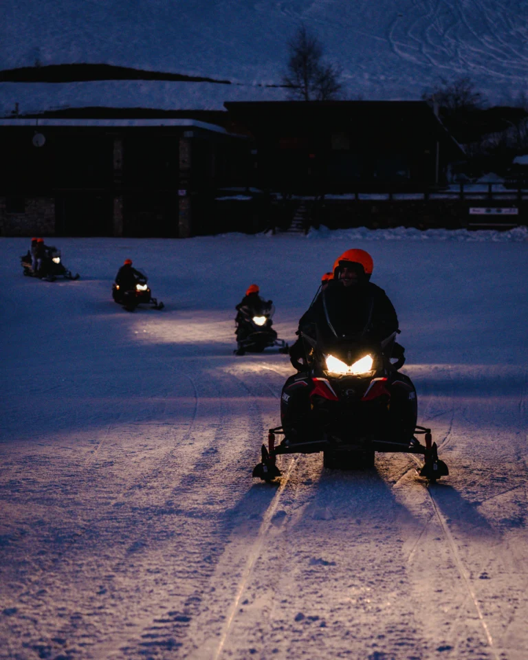 Un gruppo di motoslitte con i fari accesi percorre la neve di Prato Nevoso al calare della sera.