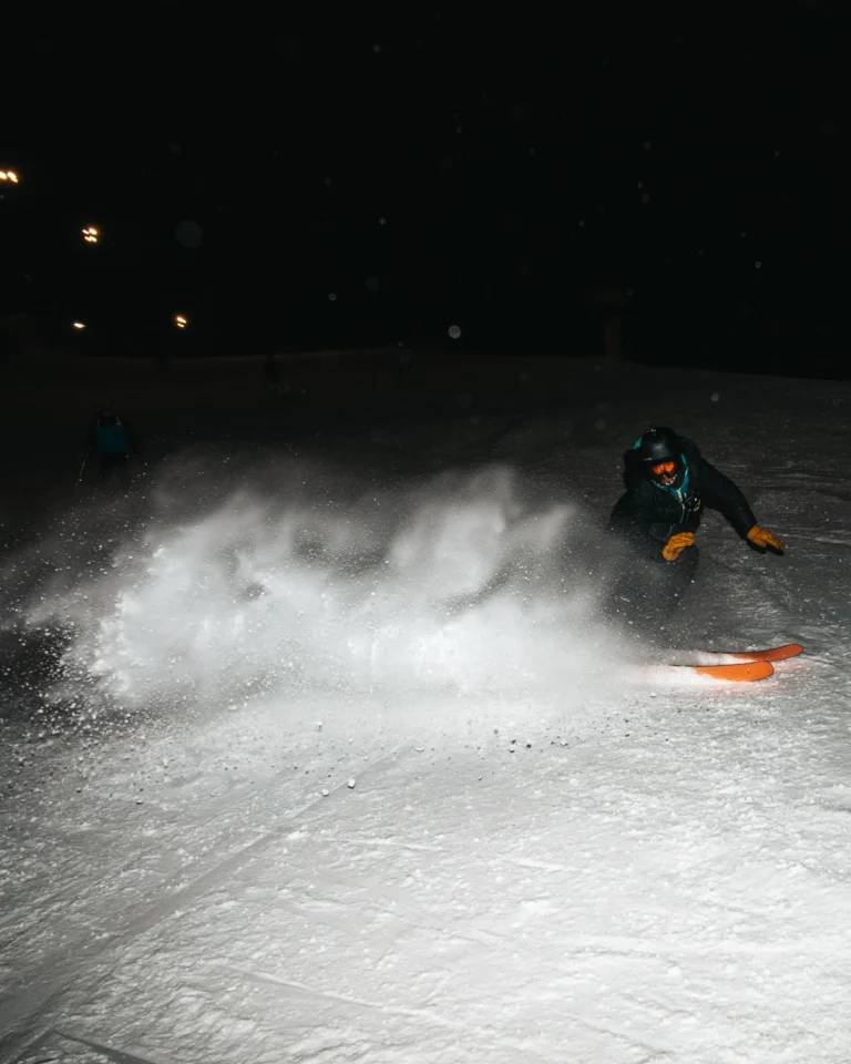 Sciatore in curva sulle piste illuminate, con sci arancioni e neve sollevata.