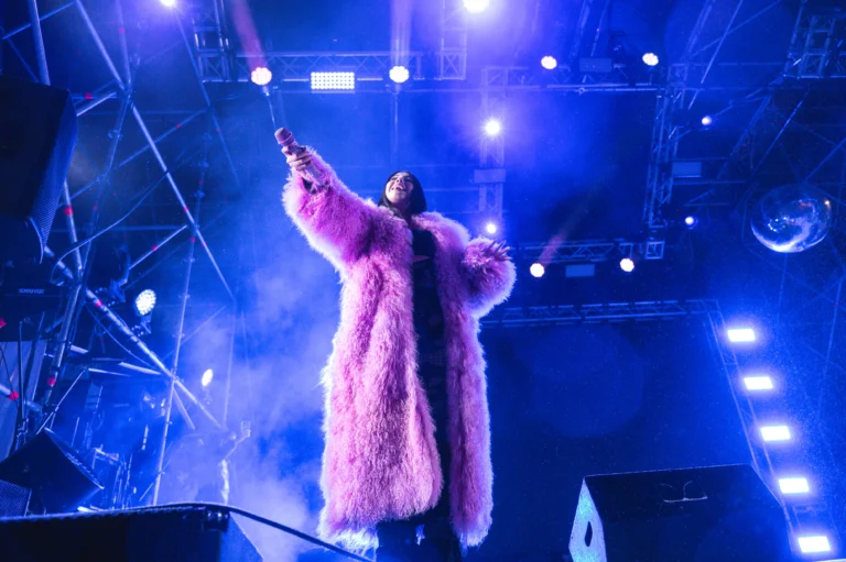 Anna Pepe sul palco alza il microfono, fari blu e cappotto rosa.