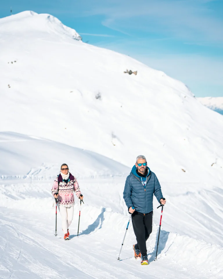 Due persone salgono con bastoncini sulla neve davanti al pendio bianco.