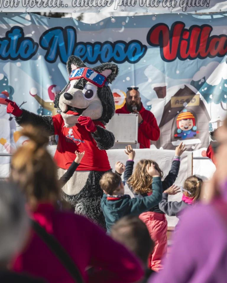 Mascotte con giacca rossa sul palco Village, con bambini che la guardano e pubblico in primo piano.