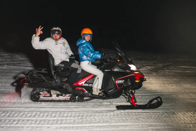 Due ospiti sorridenti con casco a bordo di una motoslitta nera e rossa sulla neve di sera.