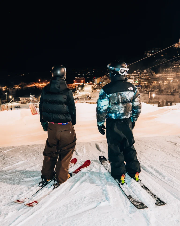 Due sciatori con casco osservano le piste illuminate di Prato Nevoso prima della discesa serale.