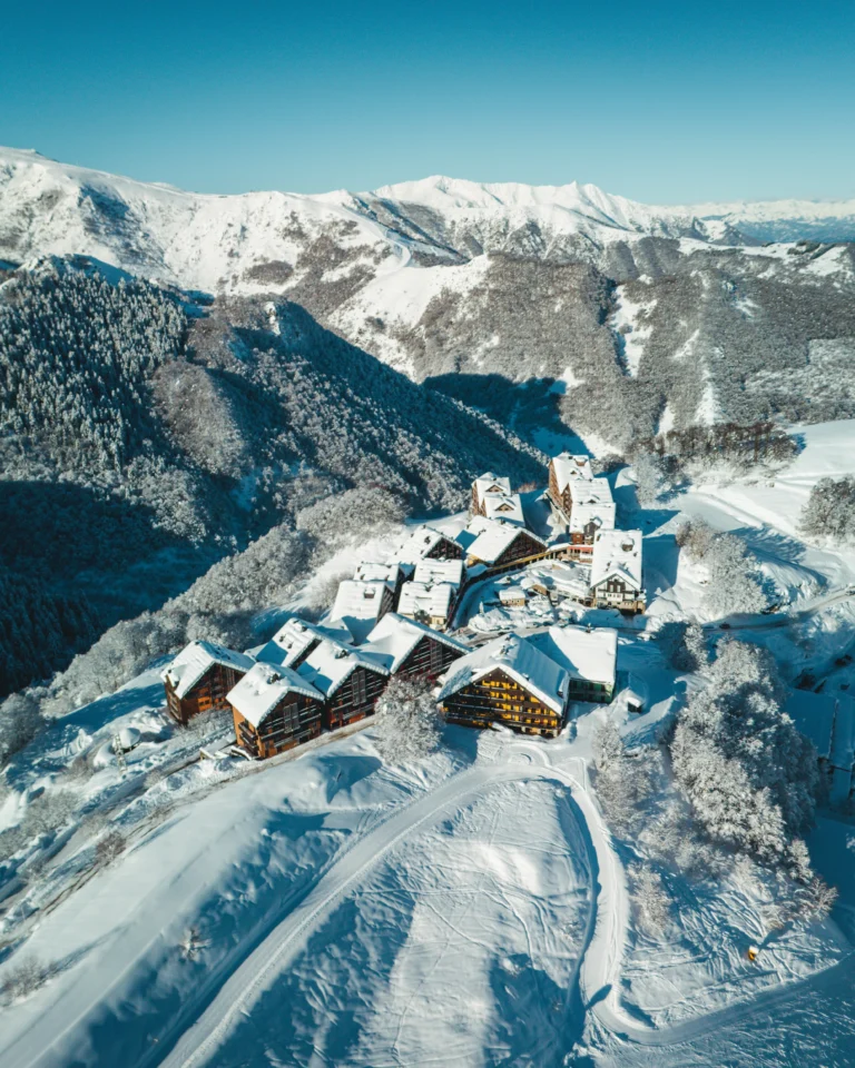 Edifici del paese innevati con montagne e bosco dietro.