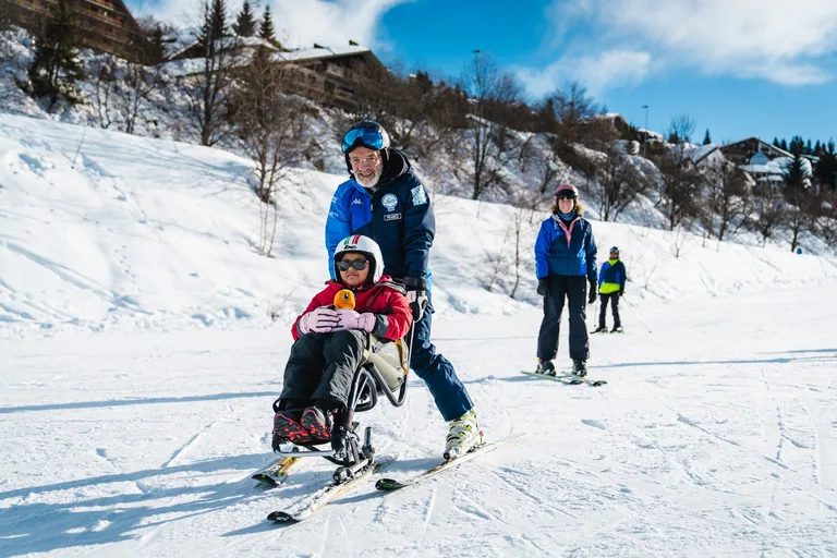 Accompagnatore guida una bambina su sci adattato, con altri sciatori sullo sfondo