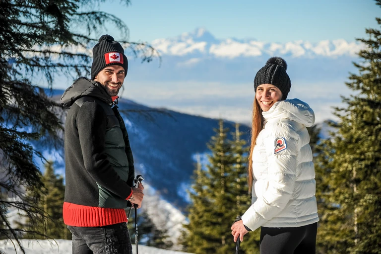 Due escursionisti sorridenti tra gli abeti, con le Alpi innevate sullo sfondo