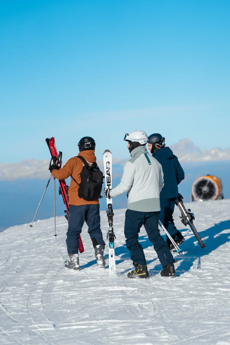 Tre sportivi con sci e snowboard sulla cresta innevata sotto cielo blu.